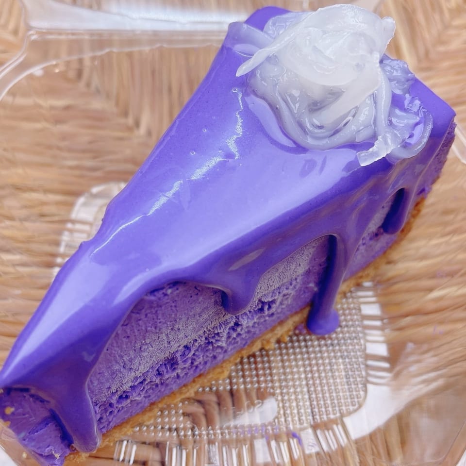 A slice of vibrant purple ube cheesecake with white coconut topping in a clear plastic container.