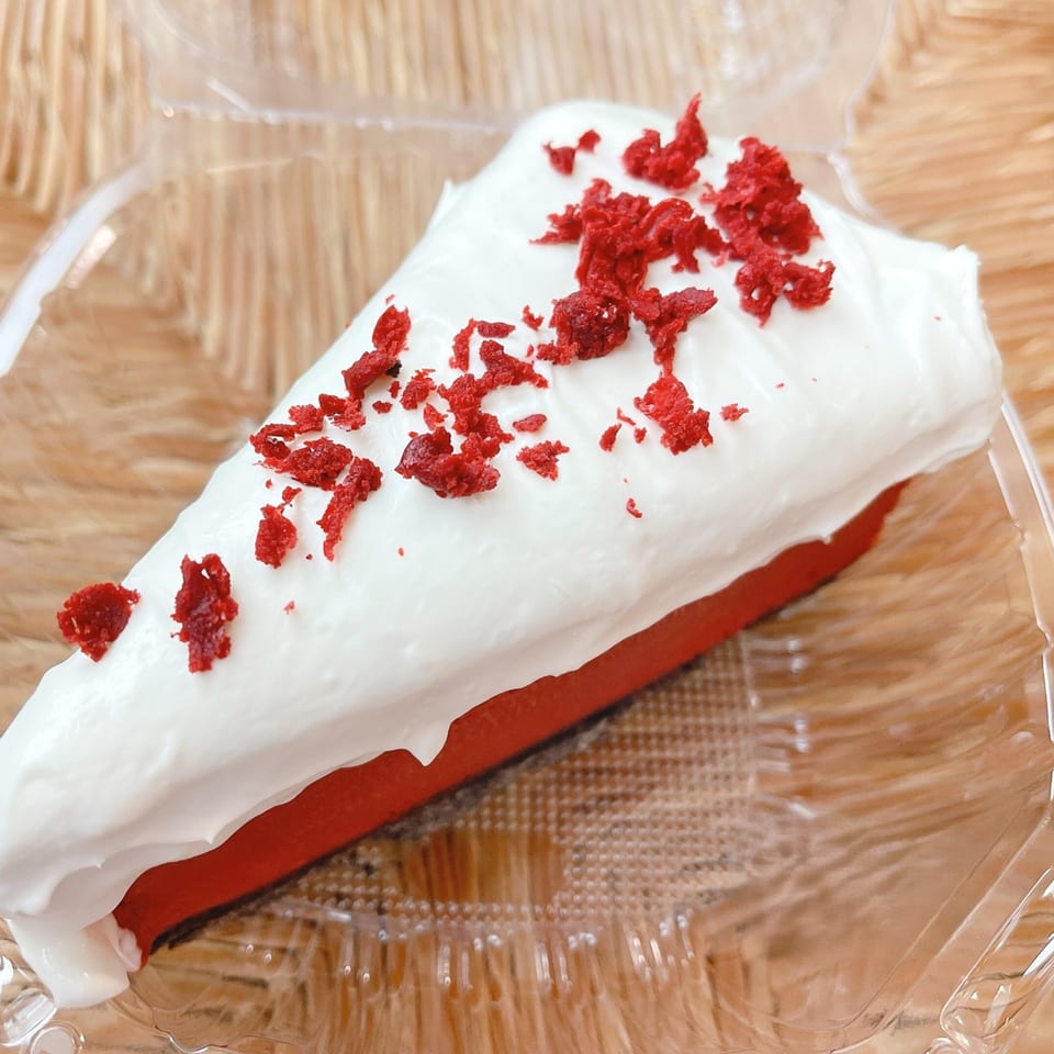 A slice of red velvet cake with white frosting and red sprinkles in a clear plastic container.