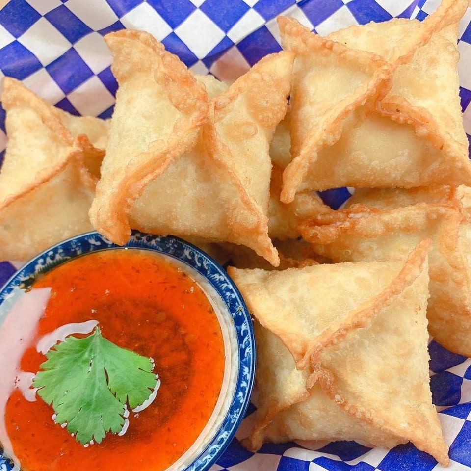 Crispy golden crab rangoons in a blue and white checkered basket with a side of sweet chili sauce.