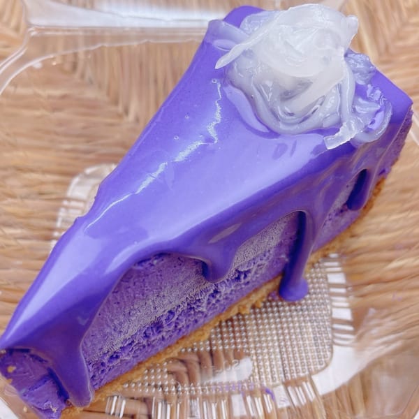 A slice of vibrant purple cheesecake with a glossy glaze and white coconut topping in a plastic container.