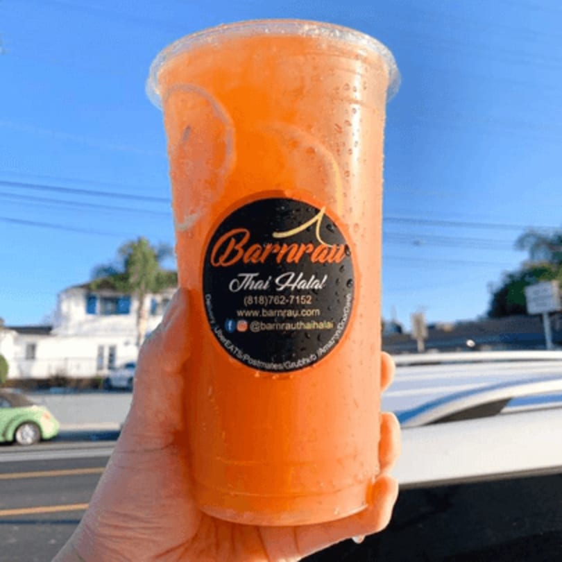 Hand holding a large plastic cup of Barnrau Thai Halal Cuisine's orange drink with citrus slices.