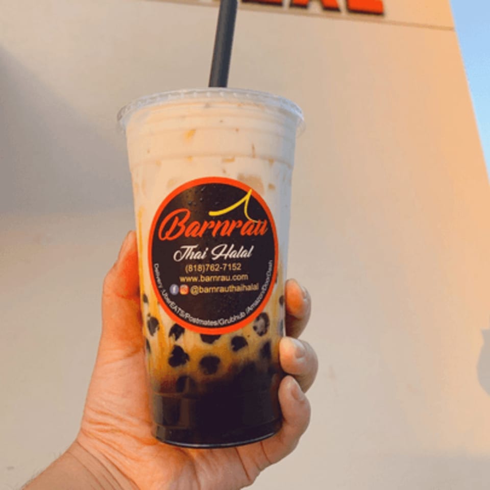 Hand holding a Barnrau Thai Halal bubble tea with tapioca pearls against a building.
