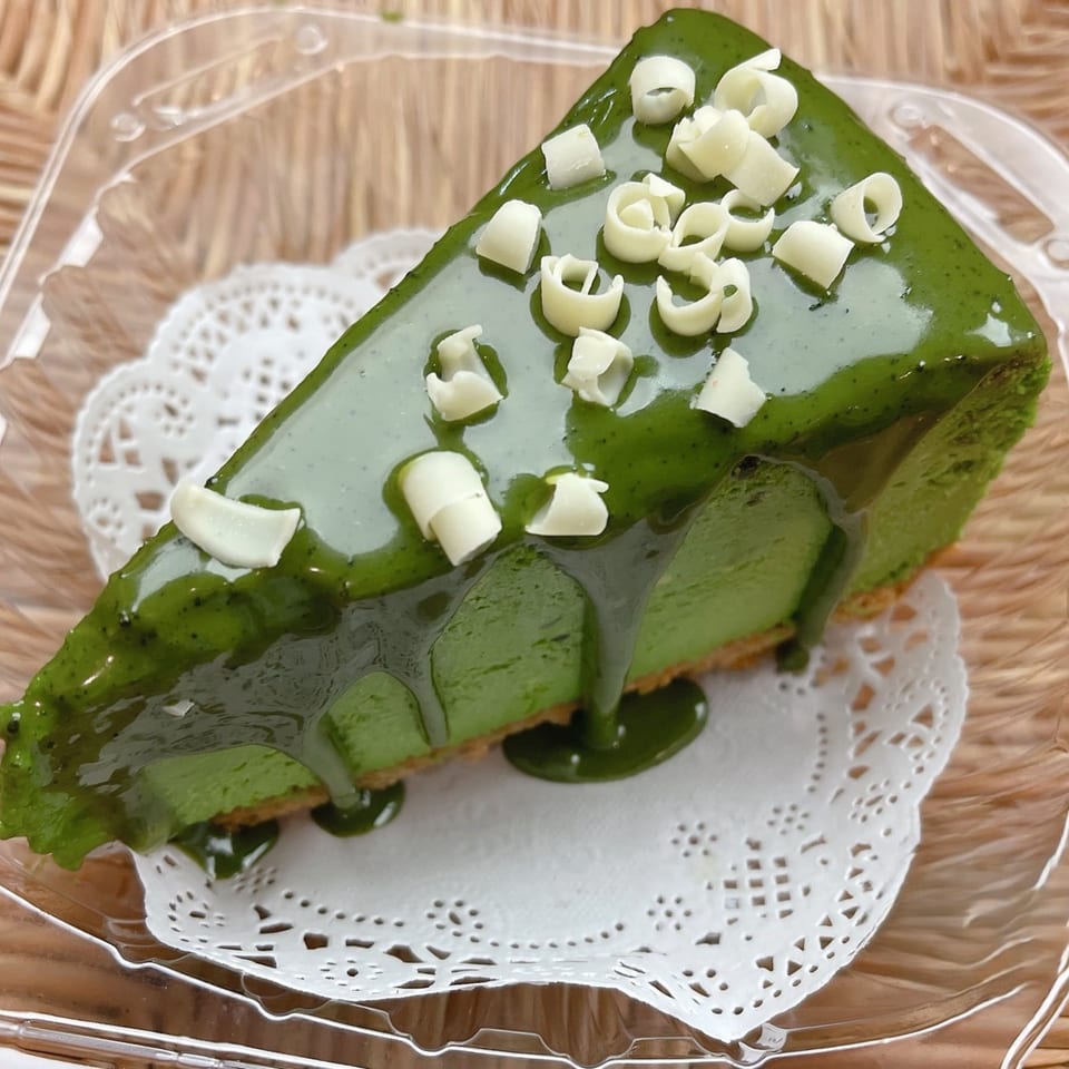 A slice of green matcha cheesecake with green glaze and white chocolate curls on a doily.
