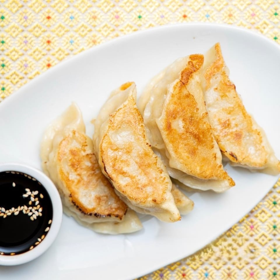 Golden-brown Thai dumplings with a side of soy sauce and sesame seeds on a white plate.