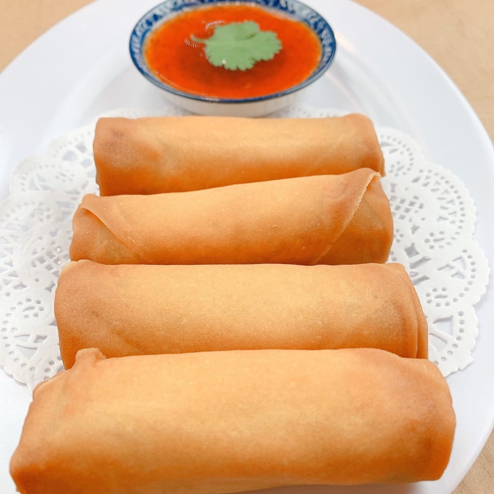 Four golden-brown Thai spring rolls on a white plate with a side of red dipping sauce.