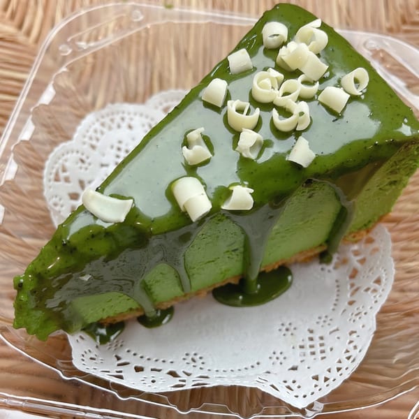 A slice of vibrant green matcha cheesecake with white chocolate curls, served in a clear plastic container.